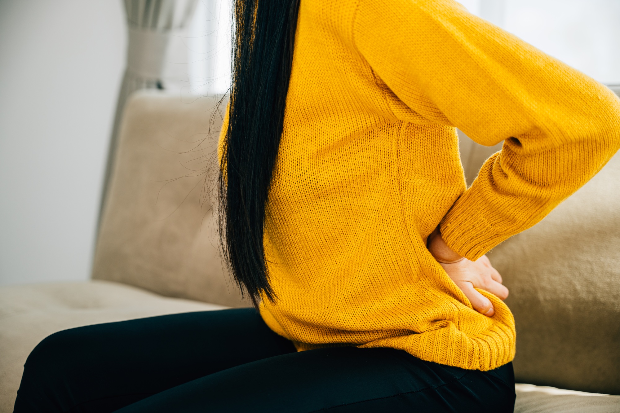 Asian woman holds her lower back in unbearable pain on a sofa